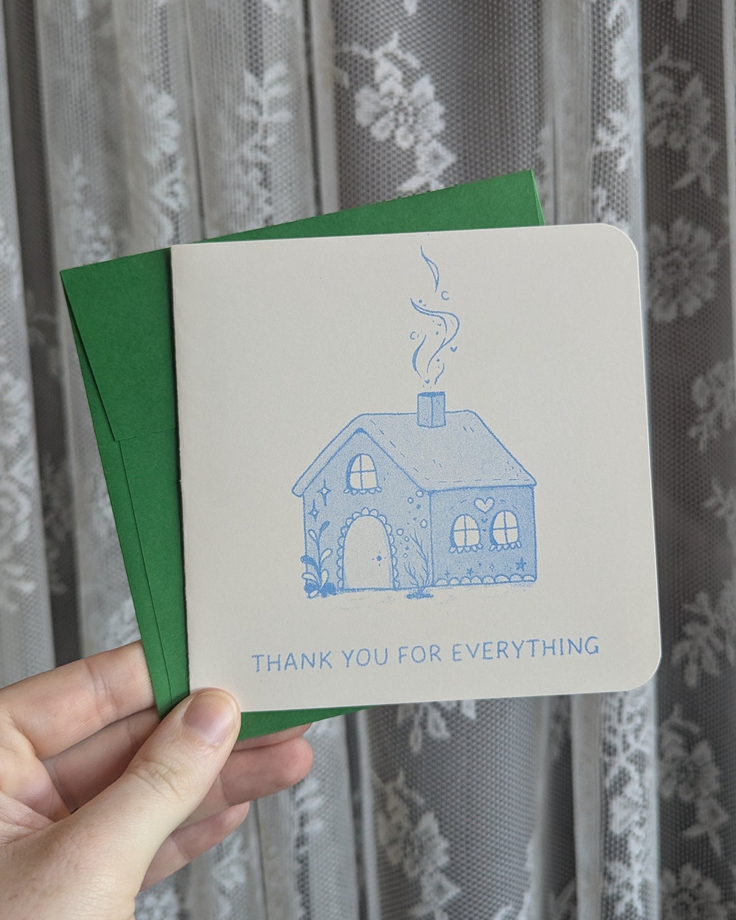 Hand holding a greeting card with a blue house illustration and text in front of a floral curtain.
