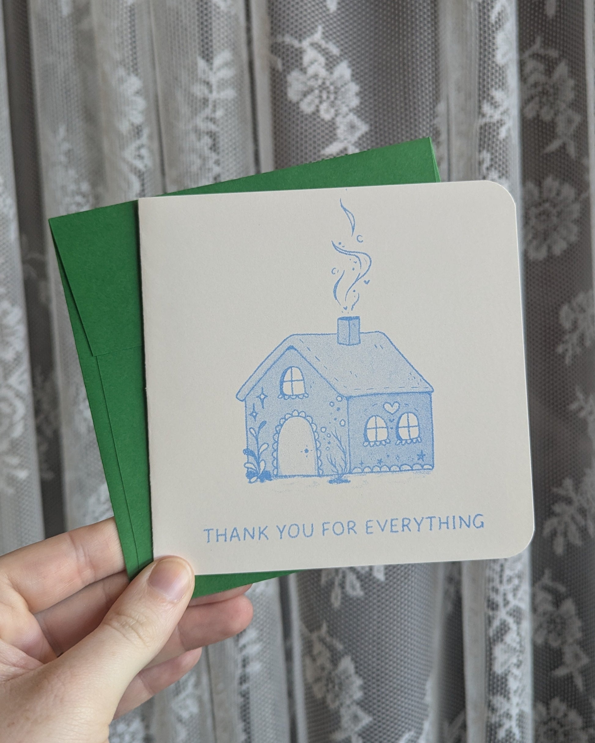 Hand holding a greeting card with a blue house illustration and text in front of a floral curtain.