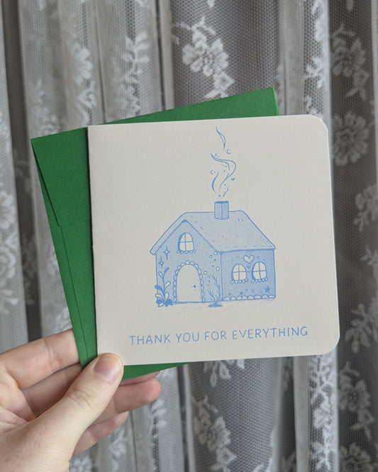 Hand holding a greeting card with a blue house illustration and text in front of a floral curtain.