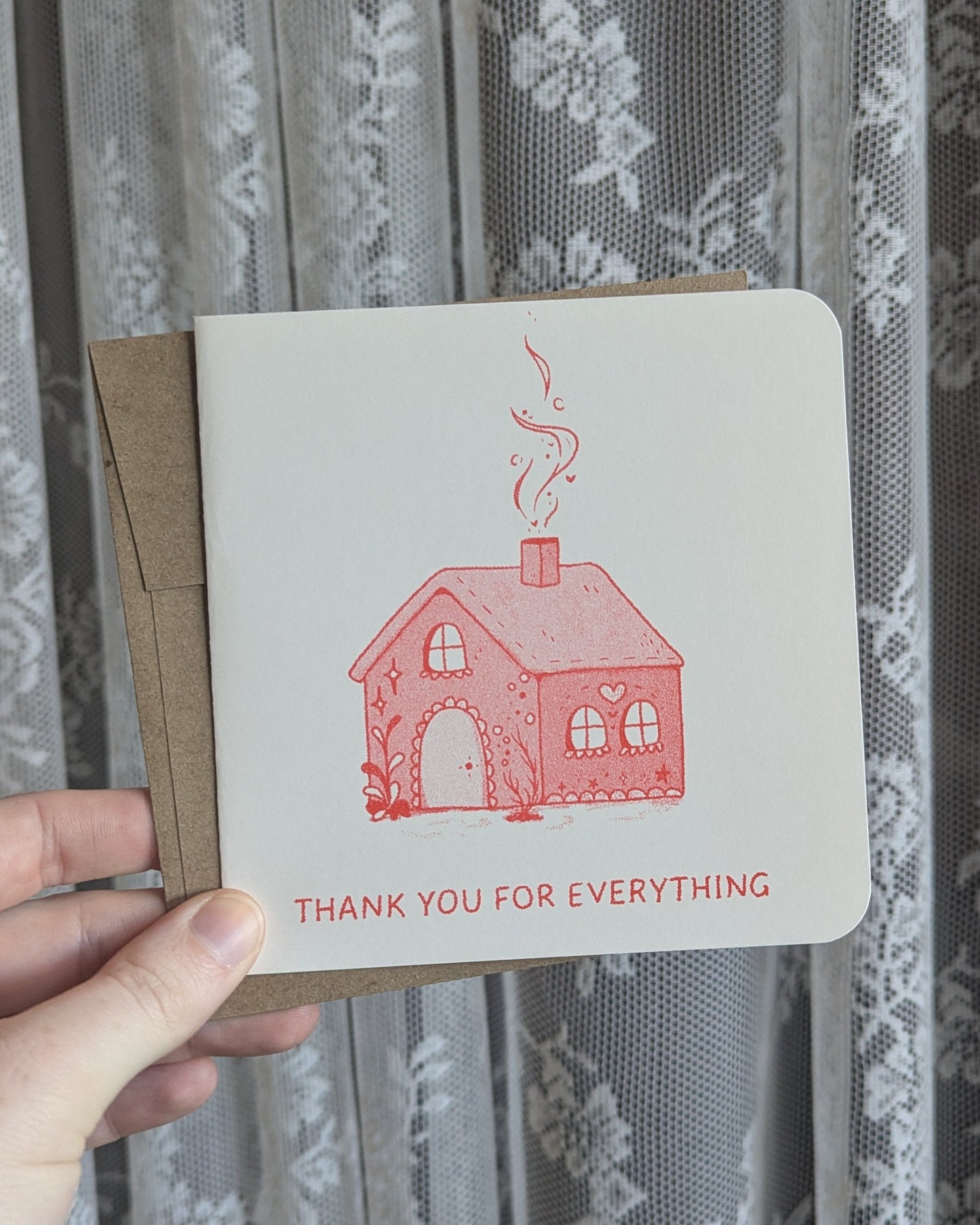 Hand holding a card with a red house illustration and text against a floral curtain background